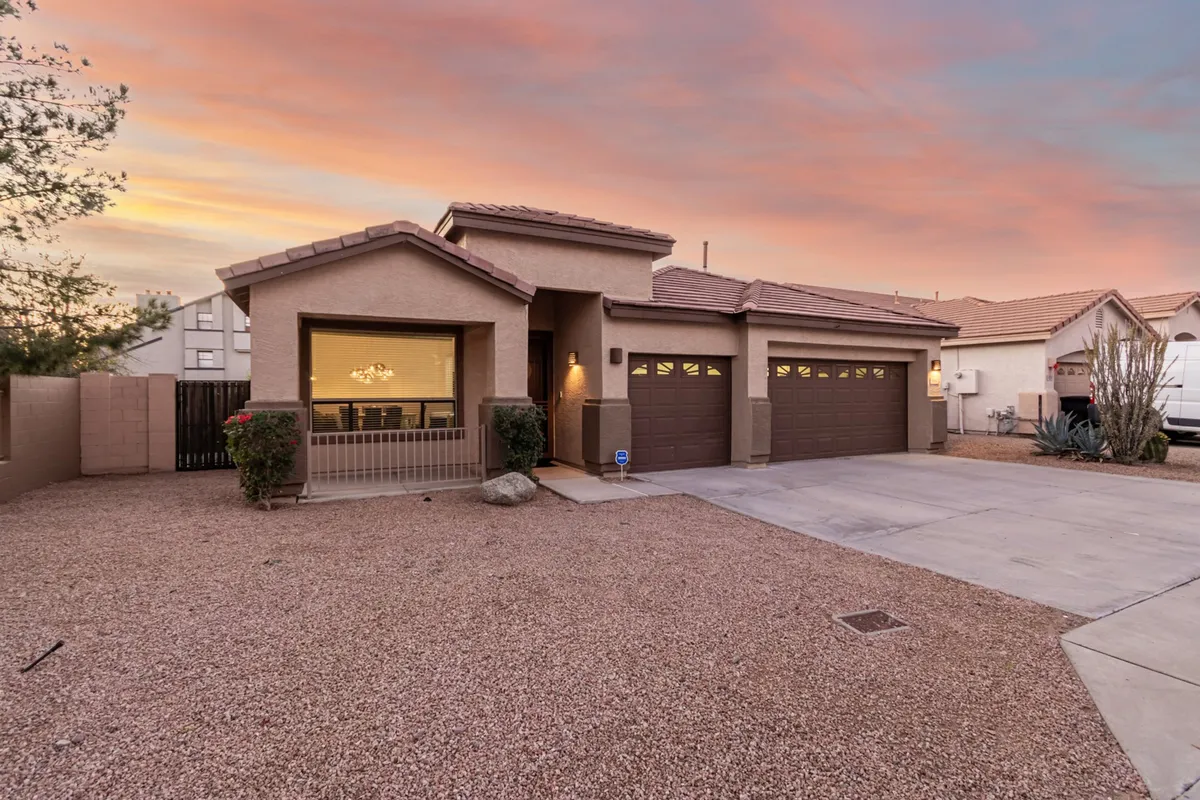 A welcoming exterior shot highlighting the front porch, prominent garage doors (including the three-car garage), and well-maintained desert landscaping. The image is taken during golden hour, casting a warm, secure glow that speaks to ease of arrival and peace of mind.