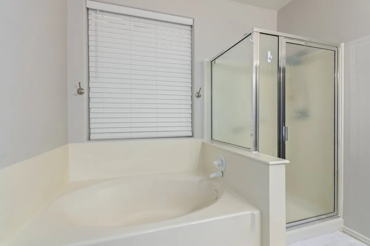 A modern bathroom with a deep soaking bathtub, a separate glass-enclosed walk-in shower, and a window with blinds. Clean lines and neutral tones evoke a tranquil, spa-like environment, making the space ideal for both relaxation and rejuvenation.