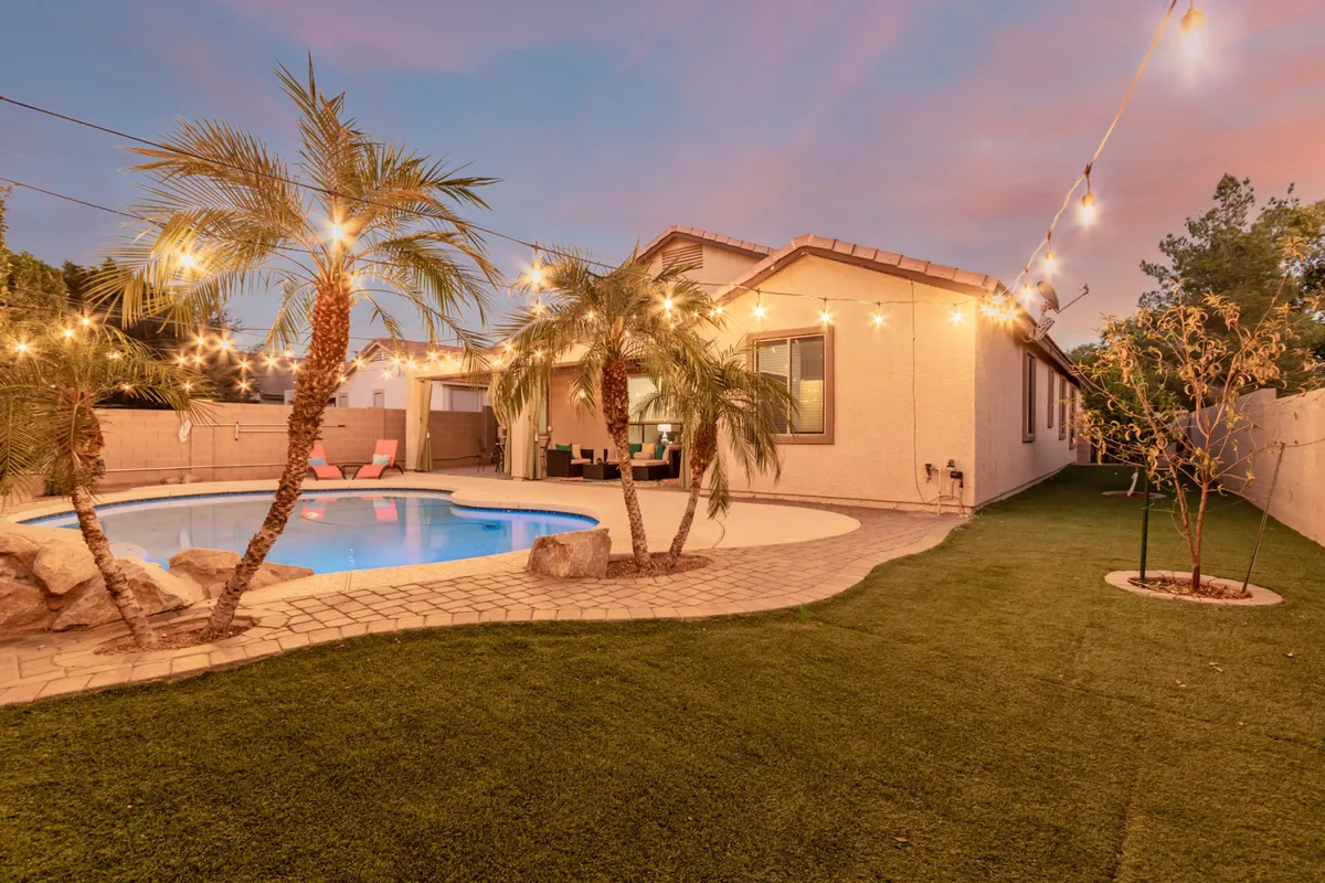A sparkling swimming pool is framed by swaying palm trees, twinkling string lights, and an inviting outdoor seating area. The warm glow suggests the perfect setting for sunset pool parties or intimate evening gatherings. The space feels festive yet relaxing, ideal for socializing and unwinding with friends after exploring Mesa's vibrant attractions.