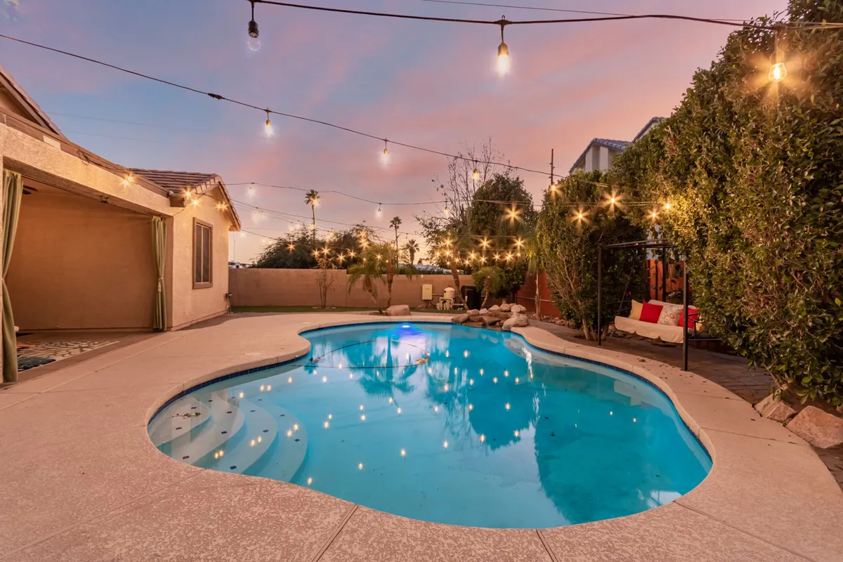 A wide exterior shot featuring a sparkling swimming pool framed by lush landscaping, sun loungers, and string lights overhead. The image includes a spacious outdoor seating area perfect for gatherings or relaxation, with hints of a BBQ setup and grassy spaces visible in the background.
