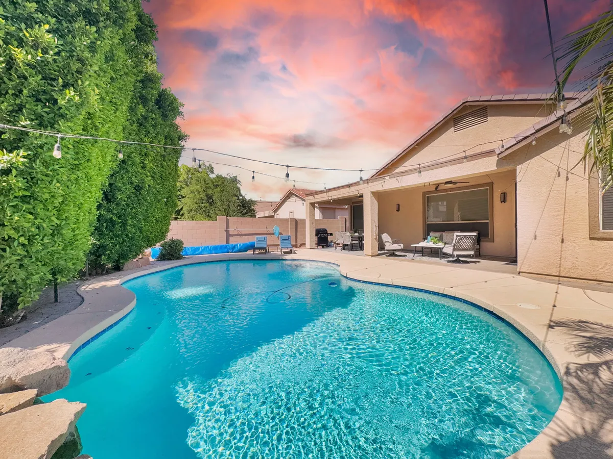 The photo showcases a serene swimming pool bordered by lush greenery and a cozy outdoor seating area. String lights twinkle above, suggesting a space ideal for both sun-filled days and intimate evenings with friends or family.