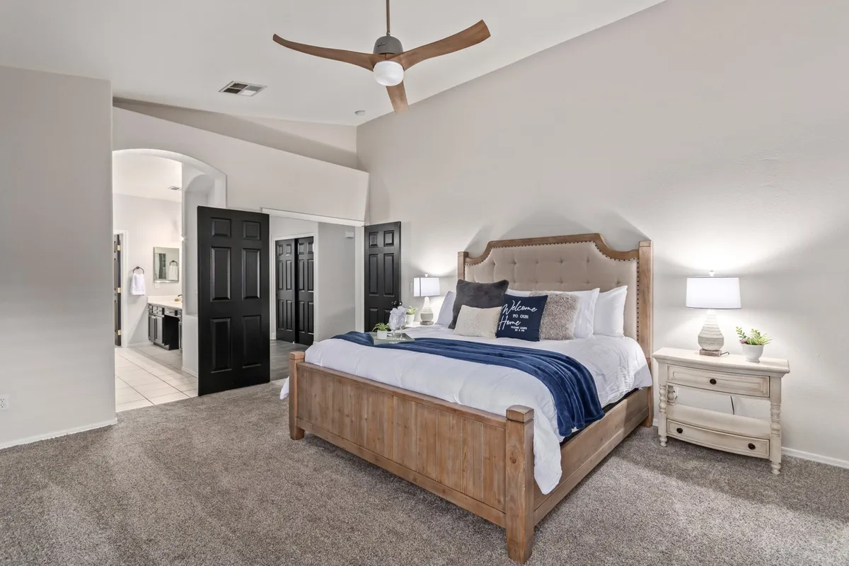 A spacious master bedroom featuring a king-size wooden bed with plush bedding and pillows, complemented by a decorative throw blanket. Nightstands with lamps flank the bed, and a modern ceiling fan hangs above. Natural light pours in from a nearby window, creating a bright and serene atmosphere ideal for relaxation.