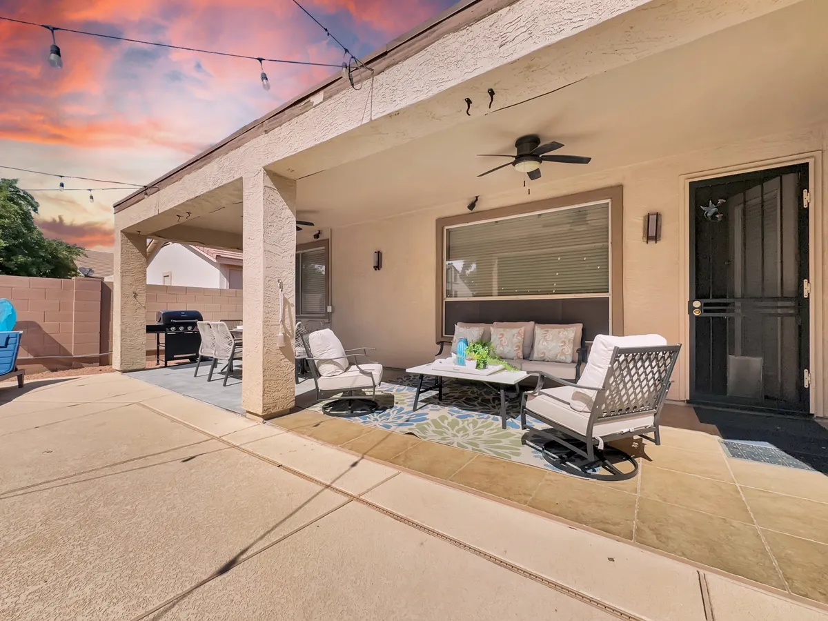 A cozy outdoor seating area is featured under glowing string lights, with a grill nearby and decorative pillows adding a touch of comfort. The scene is set for evening gatherings, morning coffees, or relaxed lounging, with the grill and covered patio clearly visible.