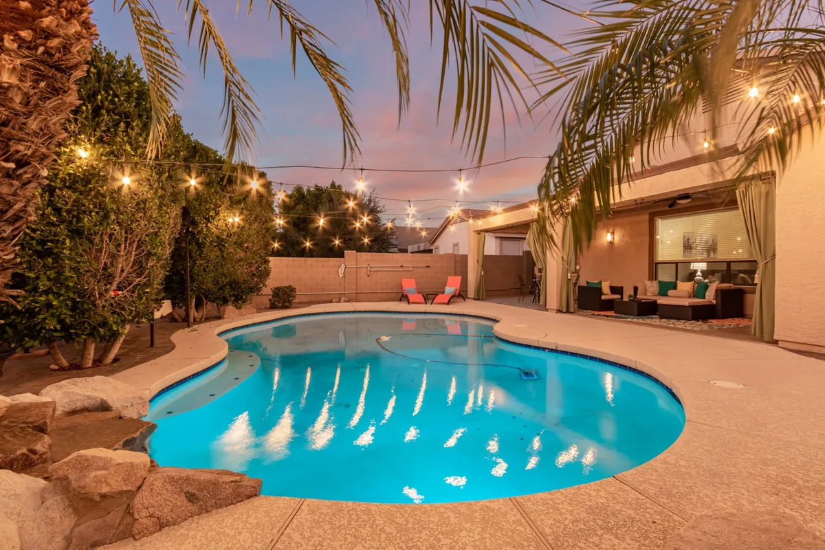 This image captures a sparkling swimming pool surrounded by lounge chairs and illuminated by hanging string lights. Palm trees offer a touch of resort-like ambiance, and the entire area exudes a welcoming, festive vibe perfect for both daytime relaxation and evening gatherings.