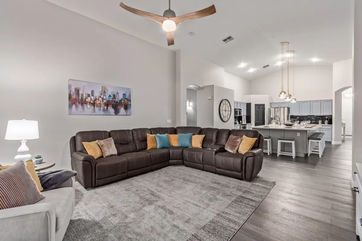 A spacious open-concept interior featuring a large sectional sofa, artwork on the wall, and a kitchen area visible in the background. The seating arrangement is perfect for family gatherings, movie nights, and socializing. The space is bright and welcoming, designed for both comfort and connection.