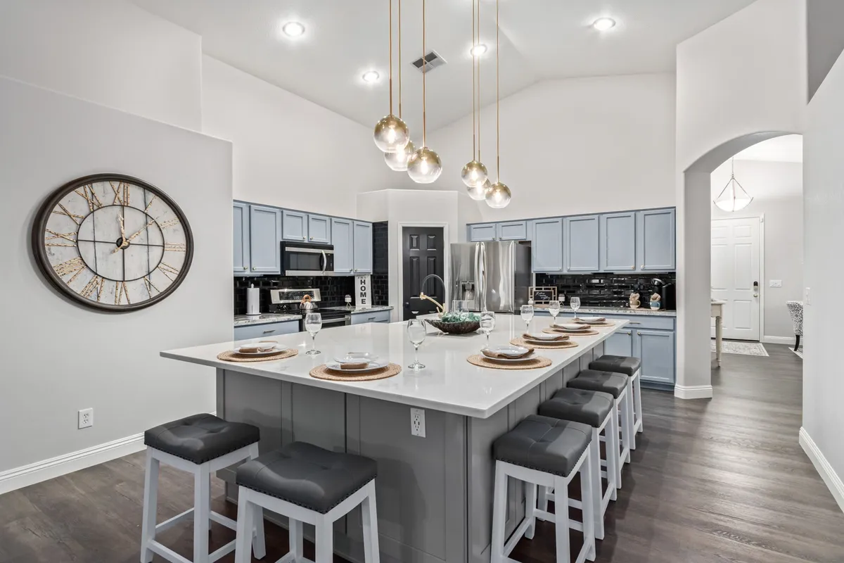 A modern kitchen highlighted by a massive island with pendant lights overhead, sleek appliances, and a wall clock. The inviting island offers ample space for meal prep, dining, and entertaining, making it a focal point for family activities.